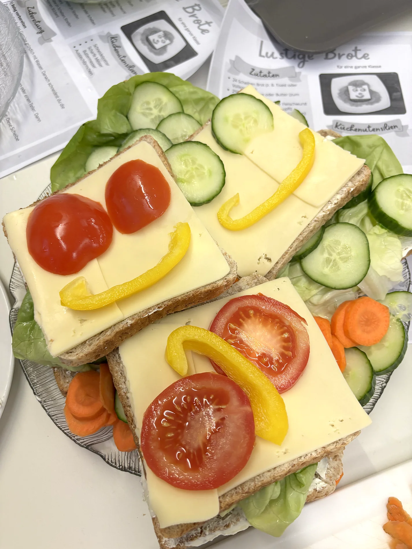 Bunte belegte Brote mit Käse, Tomaten und Paprika in Gesichter-Form angerichtet, daneben Gurken- und Möhrenscheiben.
