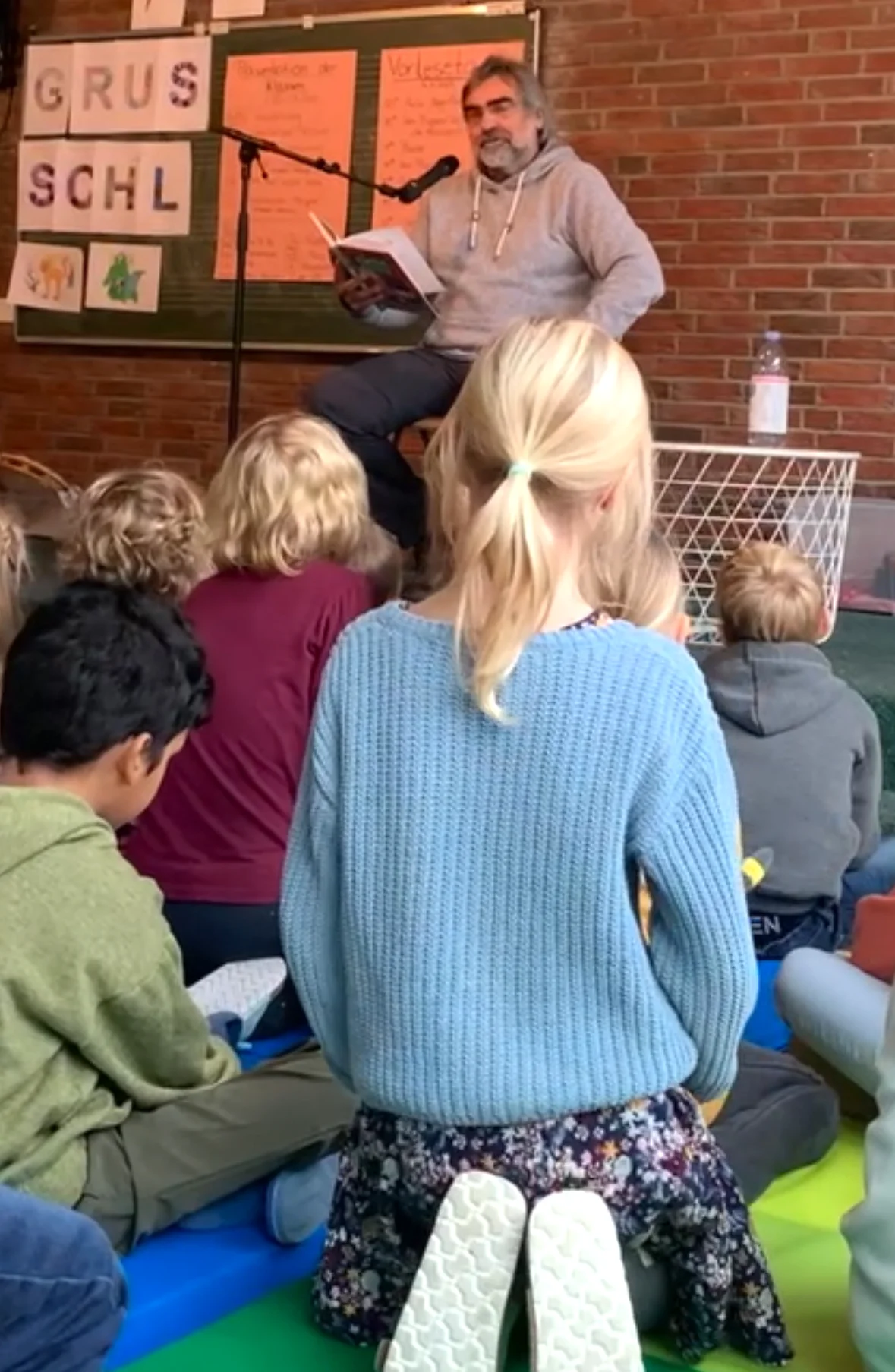 Ein Mann liest Kindern aus einem Buch vor, während diese auf bunten Matten vor ihm sitzen; im Hintergrund hängen Plakate und Buchstaben an einer Tafel.