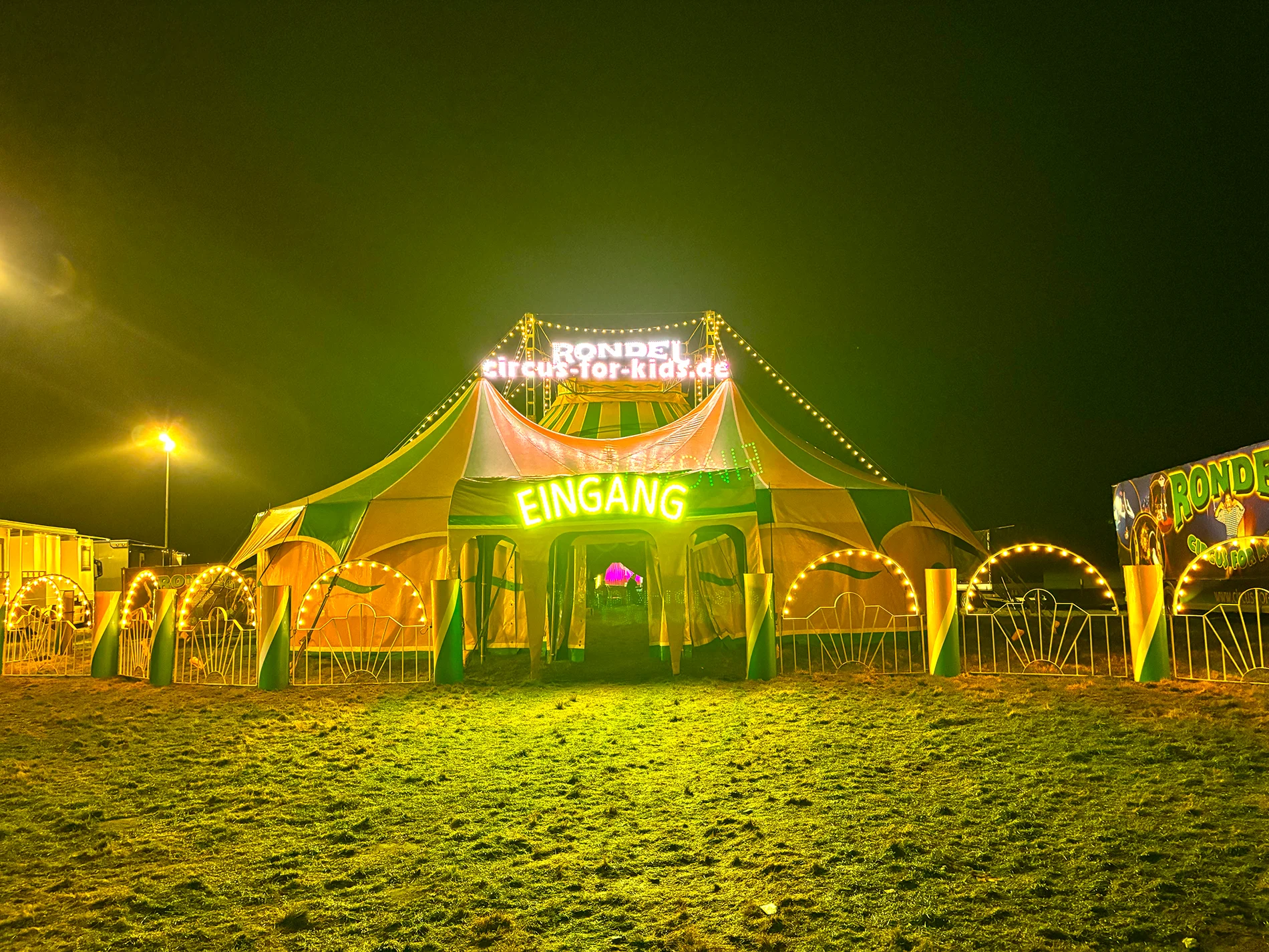 Zirkuszelt bei Nacht mit bunten Lichtern und großem „Eingang“-Schild.