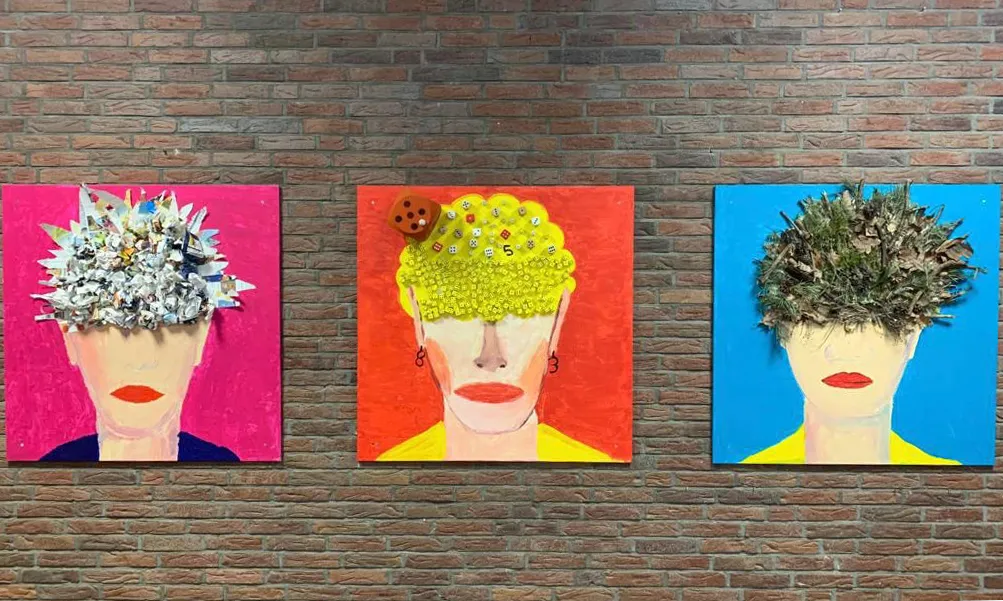 Drei farbenfrohe Collagen zeigen Porträts mit kreativ gestalteten Kopfbedeckungen aus Papier, Spielwürfeln und Naturmaterialien, ausgestellt an einer Backsteinwand der Schule. Drei farbenfrohe Collagen zeigen Porträts mit kreativ gestalteten Kopfbedeckungen aus Papier, Spielwürfeln und Naturmaterialien, ausgestellt an einer Backsteinwand der Schule.