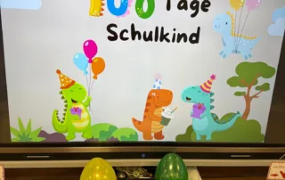 Digitale Tafel zeigt buntes Motiv‚ 100 Tage Schulkind‘ mit Luftballons und fröhlichen Dinosauriern.