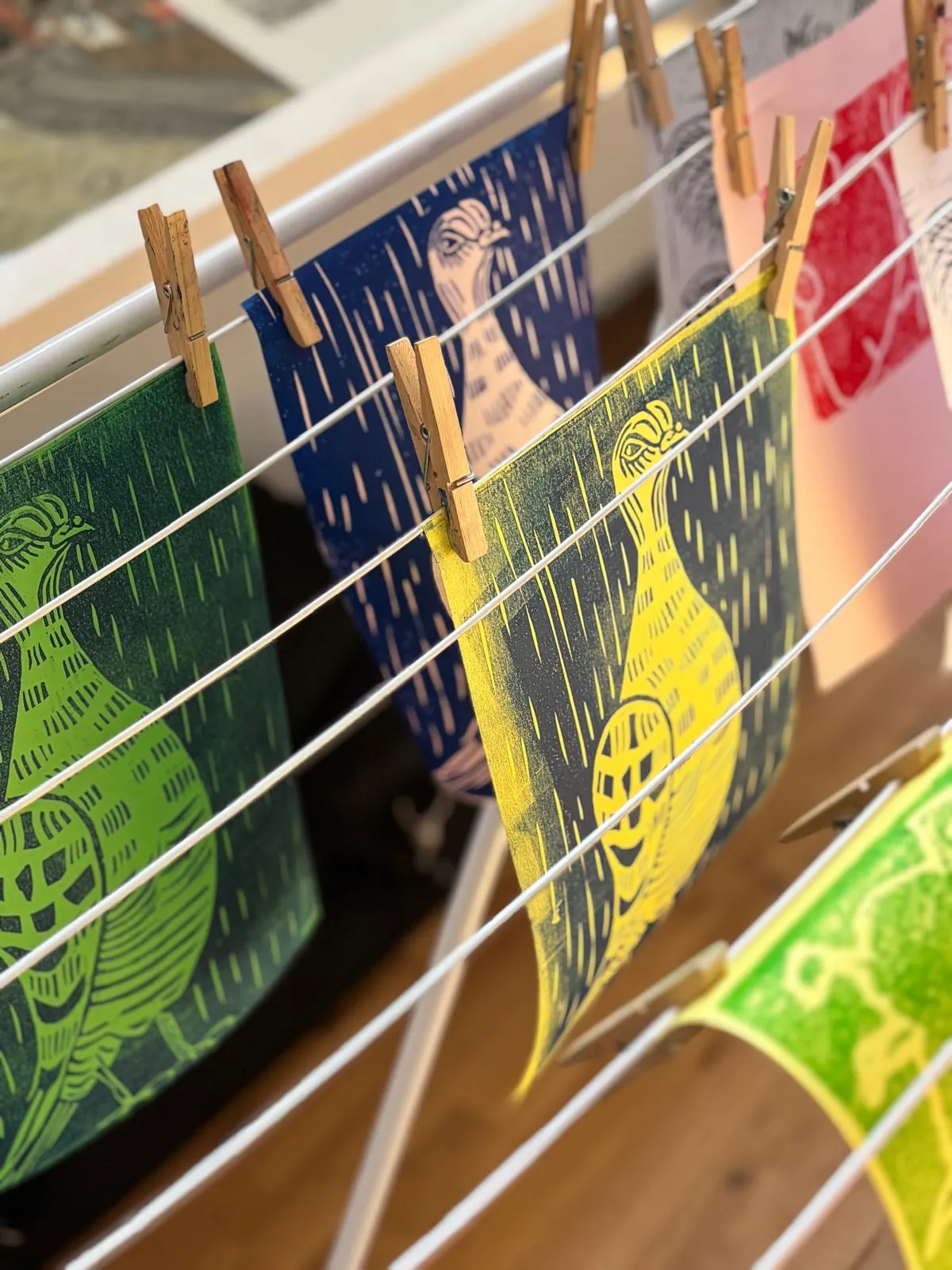Farbenfrohe Linoldrucke mit verschiedenen Motiven hängen mit Wäscheklammern an einer Leine zum Trocknen.