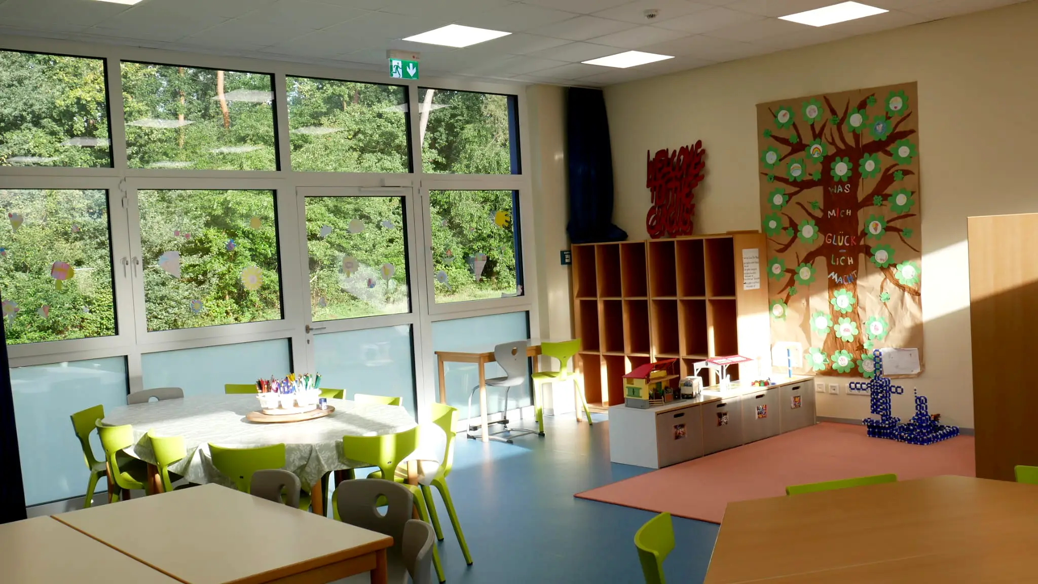ottopankockschuledrevenack_ogs1 Heller Gruppenraum mit großen Fenstern, bunten Kinderstühlen, Basteltisch, Regal und einer gestalteten Wand mit einem Glücksbaum.