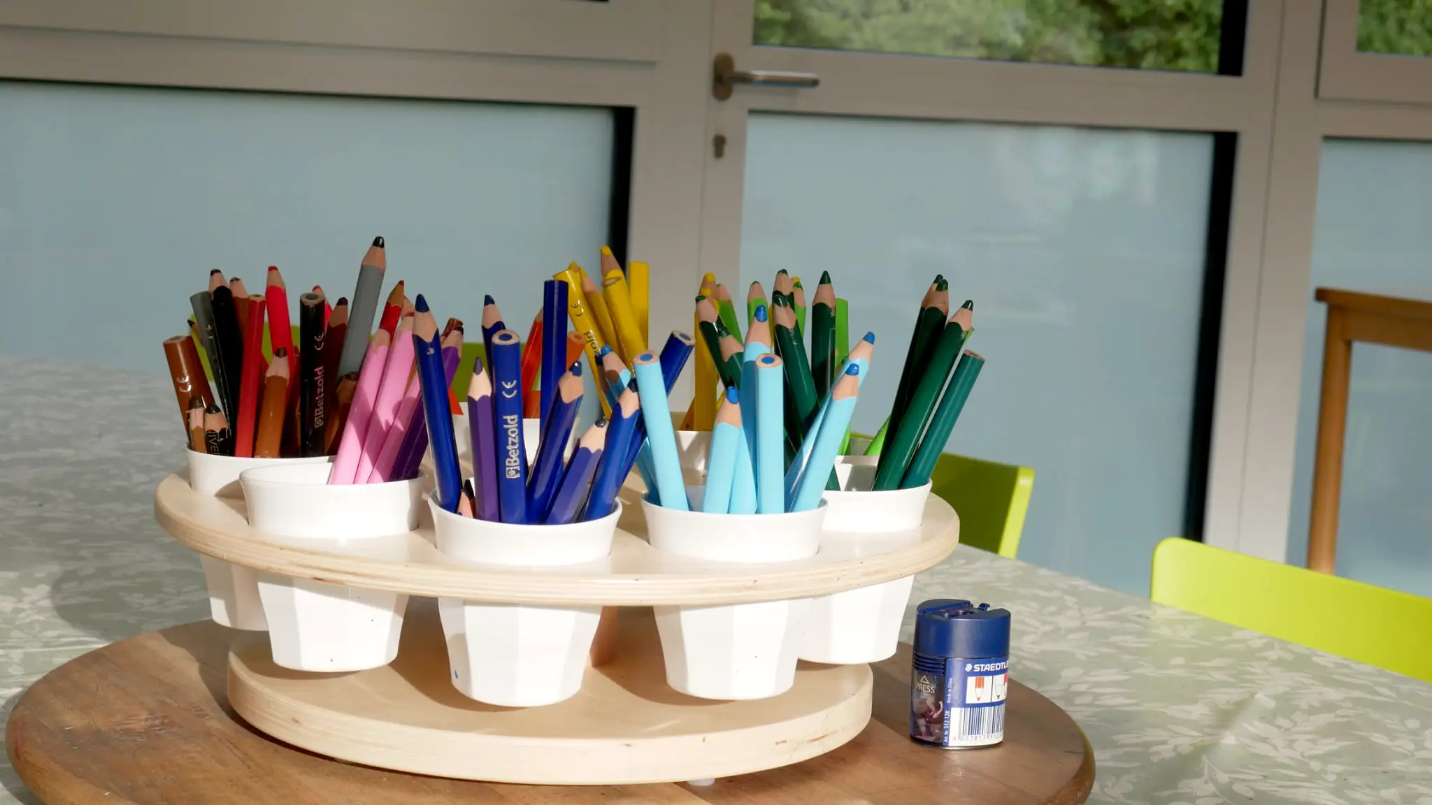ottopankockschuledrevenack_ogs3 Drehscheibe mit weißen Bechern, gefüllt mit sortierten Buntstiften in verschiedenen Farben, auf einem Tisch im Klassenraum.