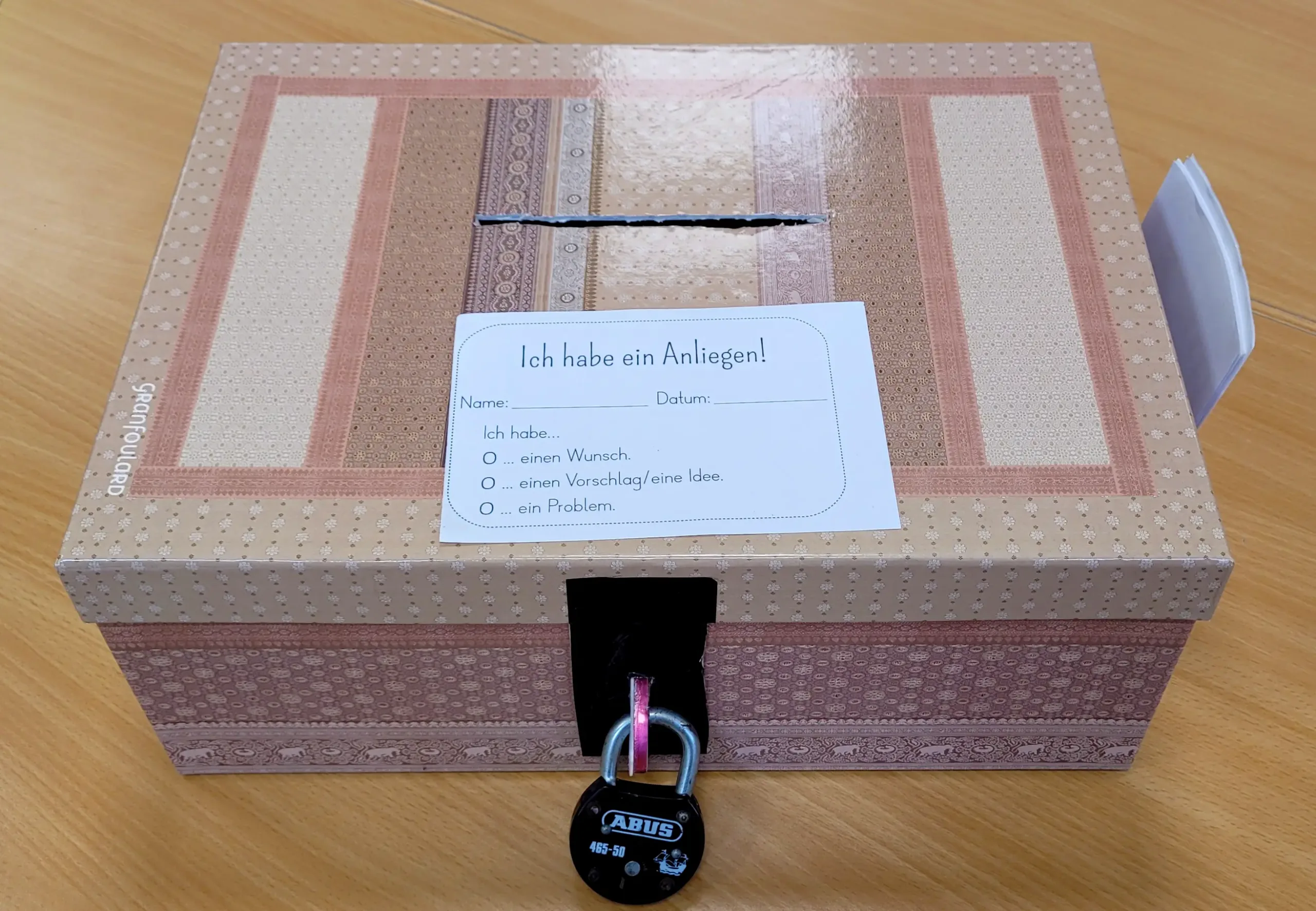 ottopankockschuledrevenack_ogsrat Dekorierte Anliegenbox mit Einwurfschlitz und Vorhängeschloss auf einem Tisch – darauf ein Zettel mit der Überschrift "Ich habe ein Anliegen!"